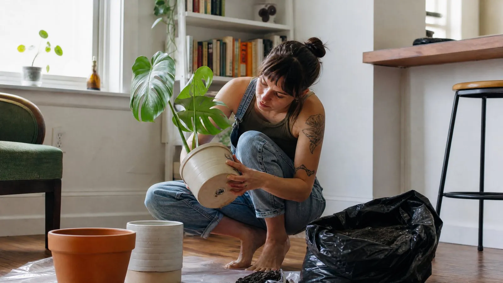 A young woman crouches on the floor of a bright apartment living room checking the drainage hole on a newly bought monstera, with empty pots and an unlabeled bag of potting soil beside her in natural window light.